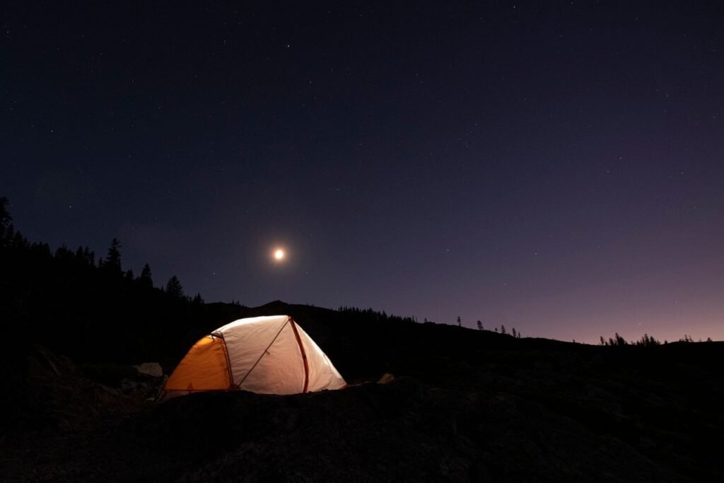 A serene night camping scene with a glowing tent under a star-filled sky and crescent moon.